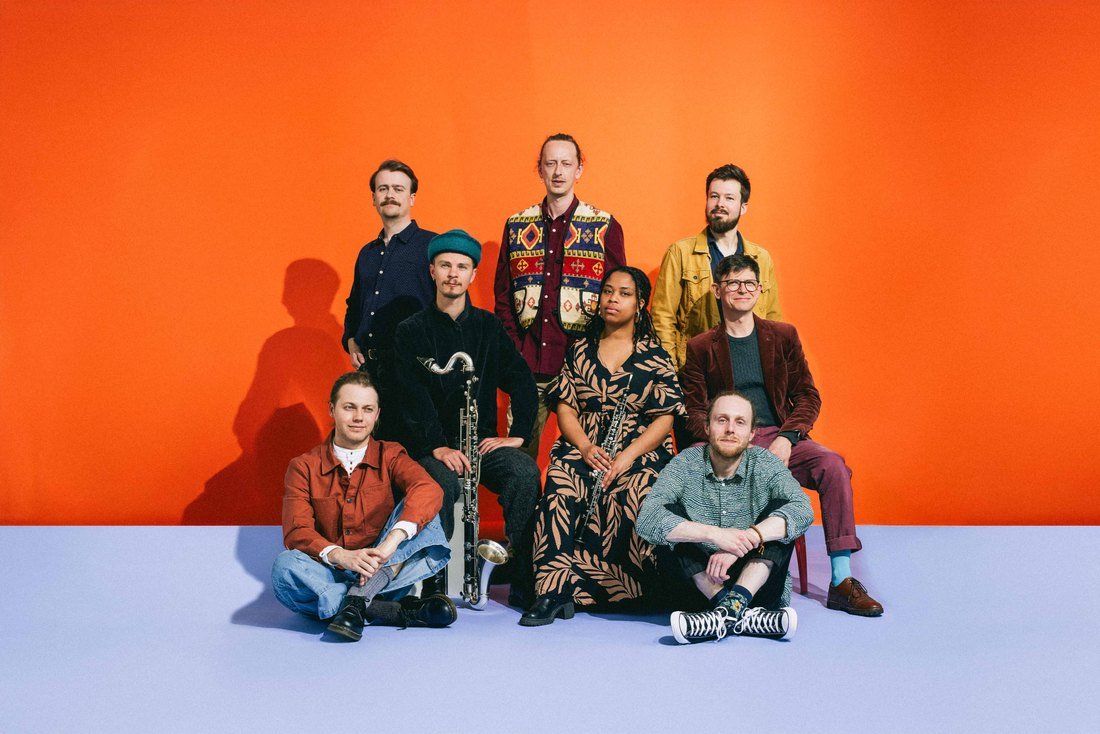 Aštuoni Ancient Infinity Orchestra nariai studijoje ryškiai oranžiniame fone ant šviesiai alyvinių grindų, tarp jų stačias didelis pučiamasis instrumentas