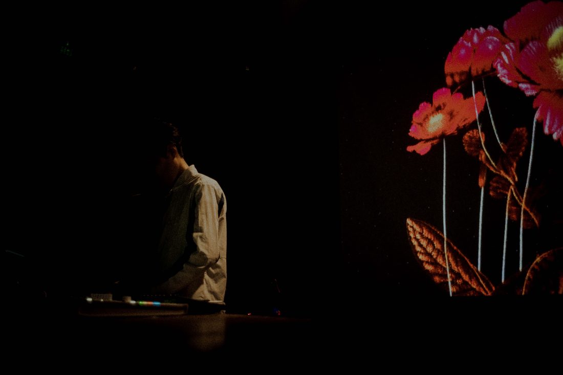 aus bei einem Live-Auftritt, über seine Instrumente gebeugt, neben einer Projektion roter Blumen