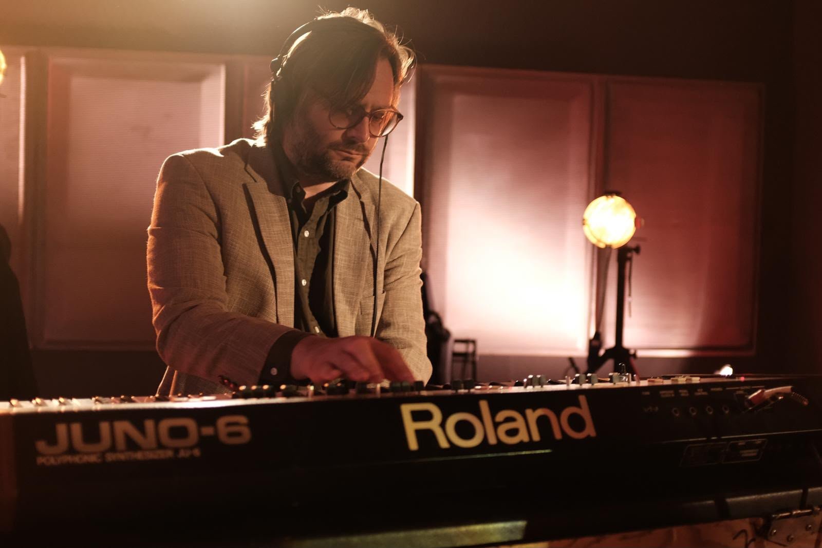 Greg Foat in headphones and a pale linen jacket, working the controls of a Roland Juno-6 synthesiser in warm studio light