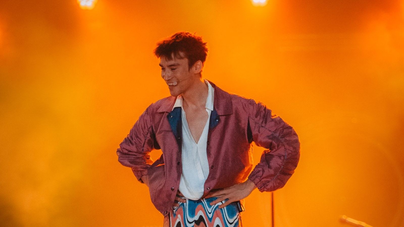 Jacob Collier on stage in orange light, hands on hips and headset microphone on, during his solo Djesse show at the Positivus Festival, 2024