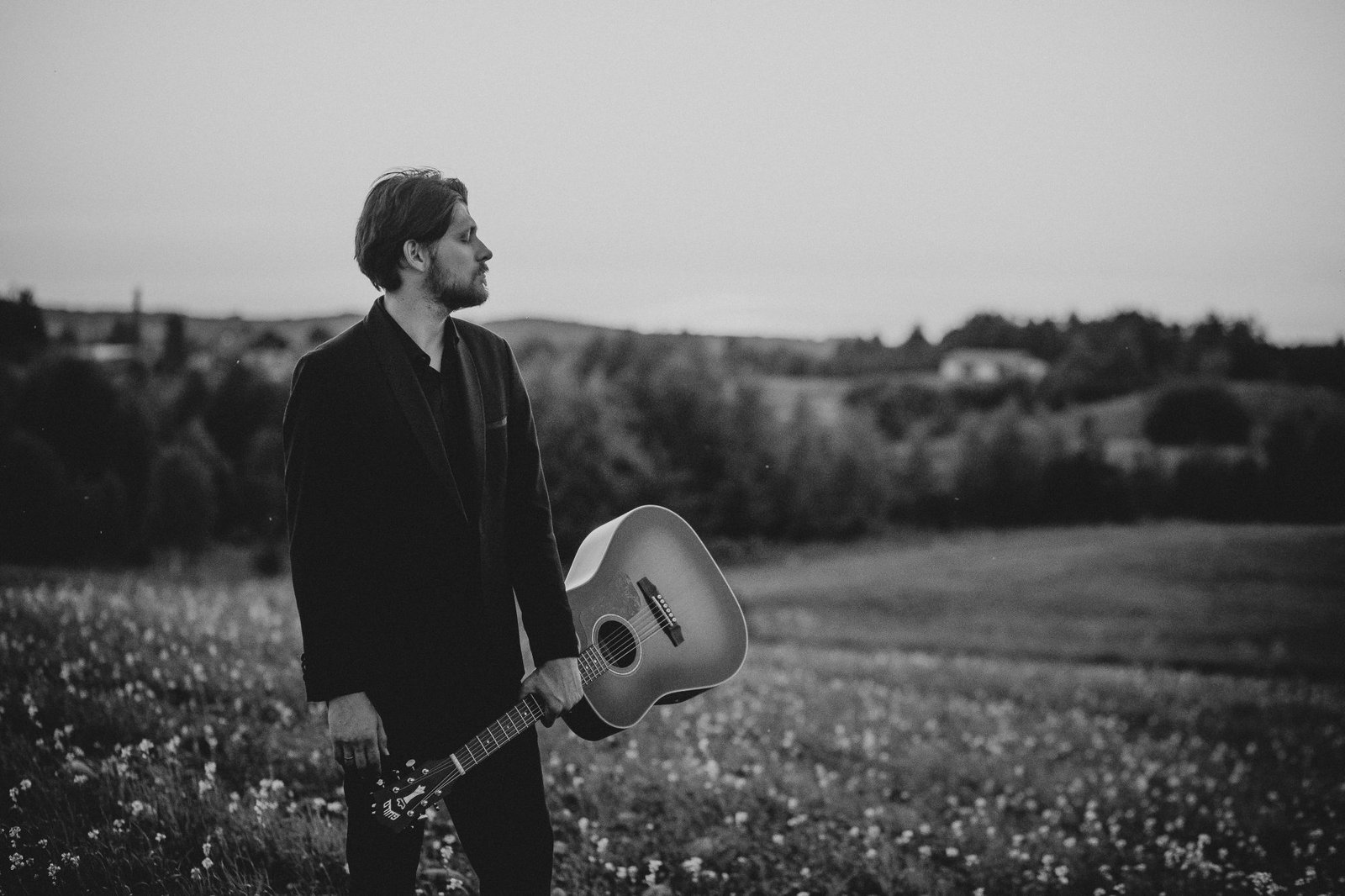 Justinas Stanislovaitis with an acoustic guitar in a Lithuanian meadow at dusk — black-and-white press portrait