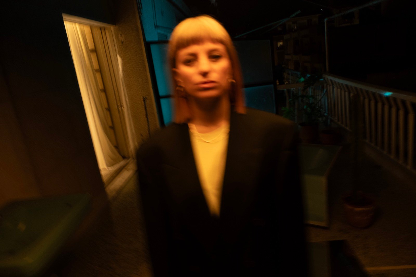 Maria Chiara Argirò at night on a balcony, facing the camera in a dark oversized blazer over a pale yellow top, her face lit warm amber and blurred by movement, a curtained doorway on the left of the frame and a metal railing and potted plants on the right