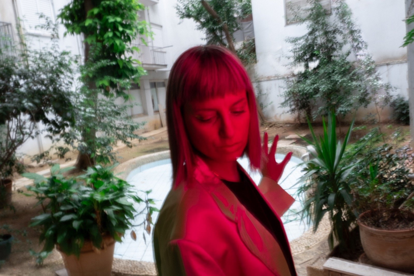 Maria Chiara Argirò standing in a green planted courtyard, eyes closed, one open hand raised beside her face, a deep crimson light falling on her alone while the courtyard behind stays daylit — motion-softened colour portrait