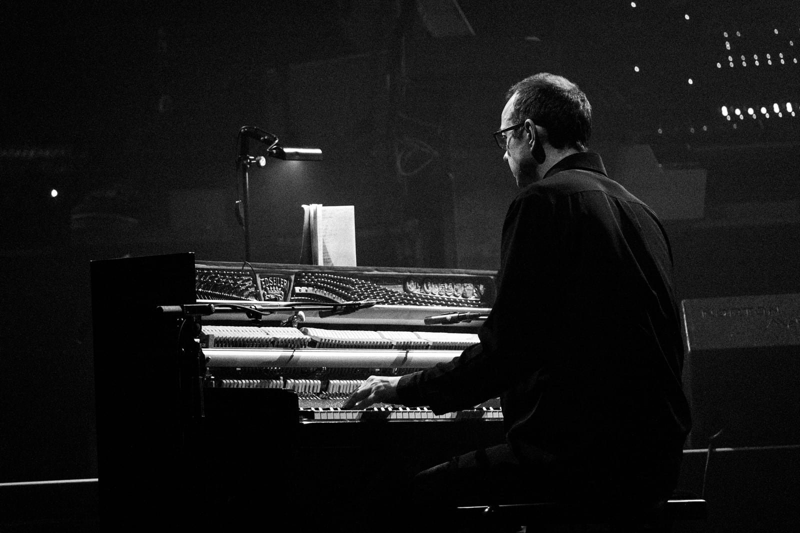 Otto A. Totland seen from behind at an upright piano with its front panel removed, the strings and hammers exposed, a clip lamp lighting a single sheet on the music desk — black-and-white stage photograph