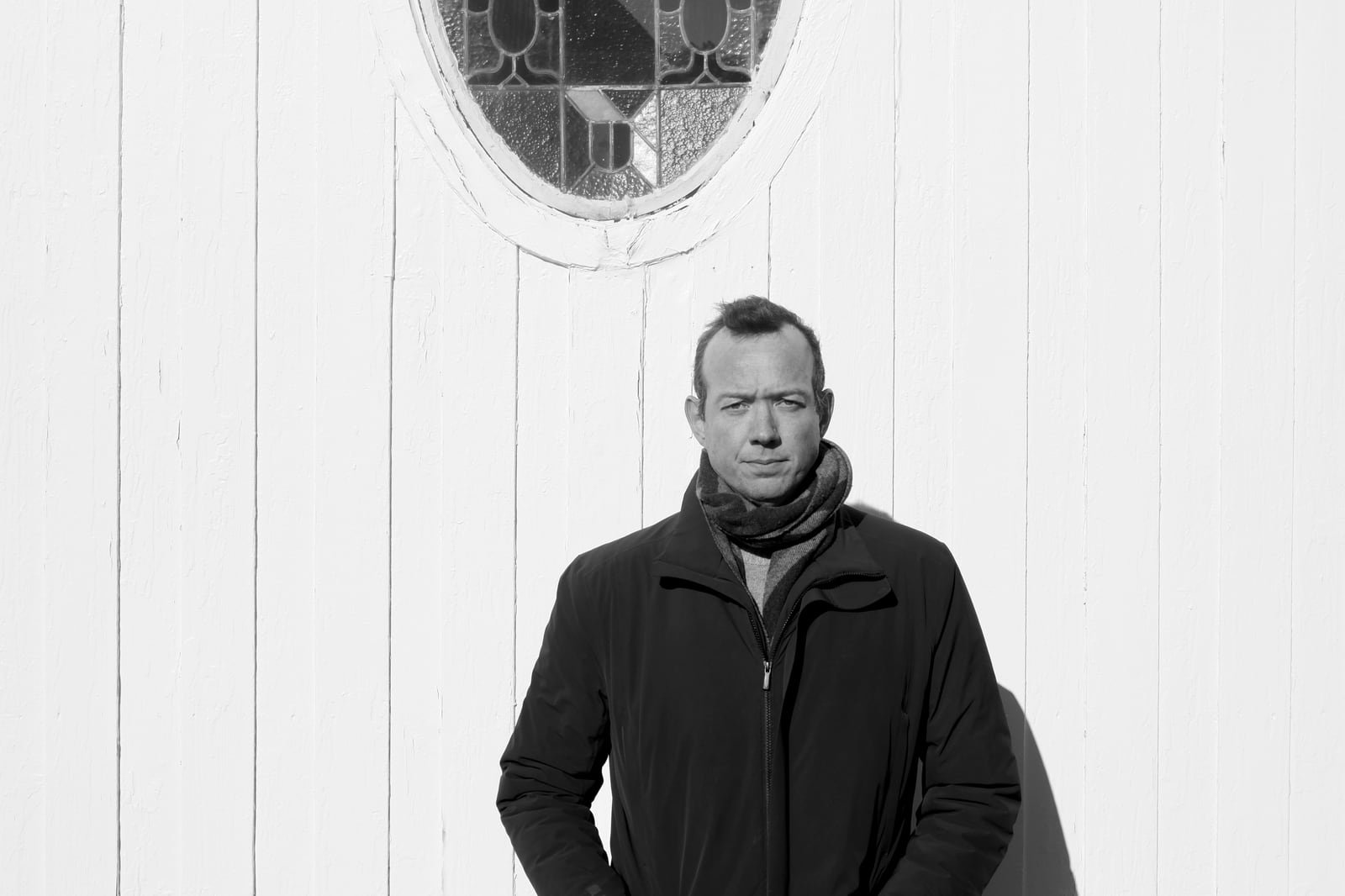 Otto A. Totland in a dark zipped coat and scarf, standing in hard low sun against a white-painted wall of vertical planks, an oval leaded-glass window above him — black-and-white portrait