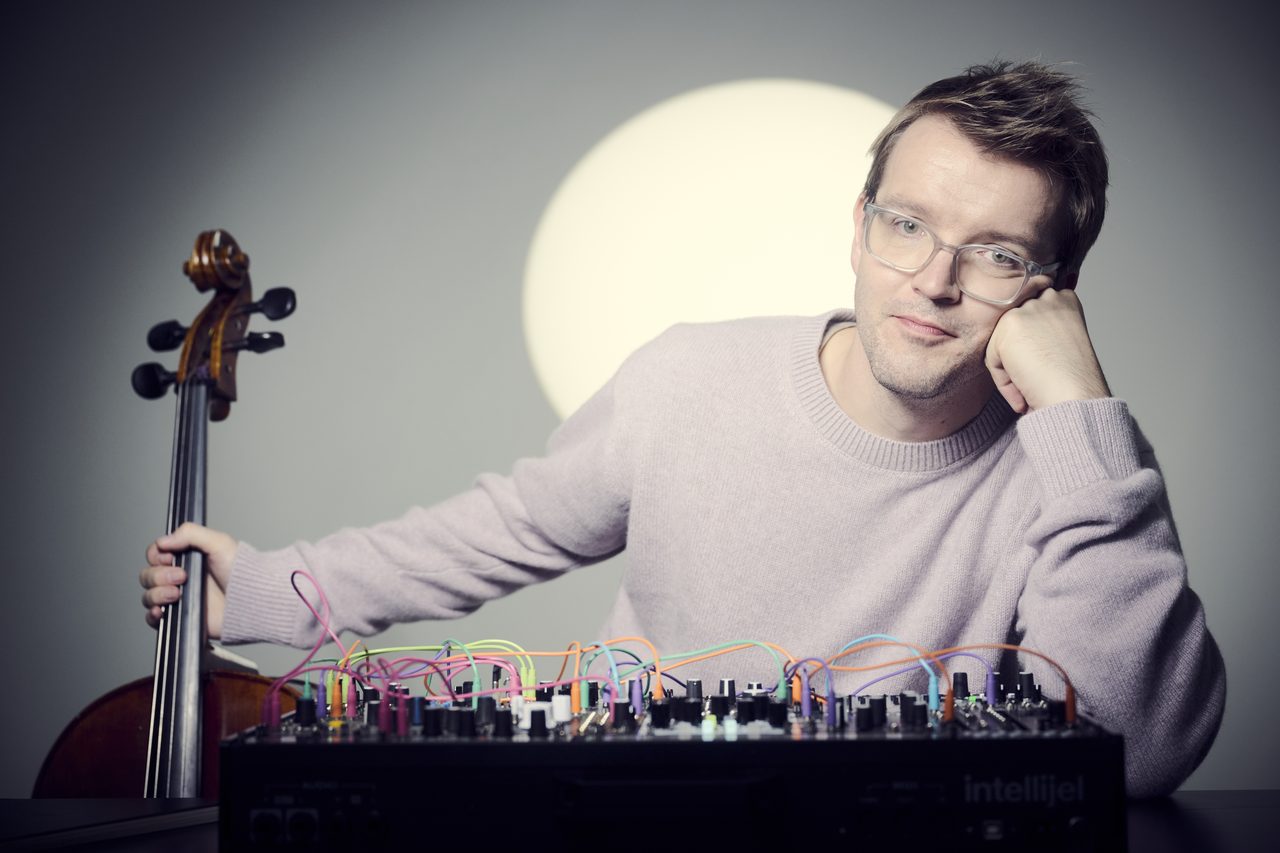Peter Gregson sitzt mit seinem Cello und einem Modularsynthesizer voller farbiger Patchkabel, hinter ihm ein Kranz aus Licht
