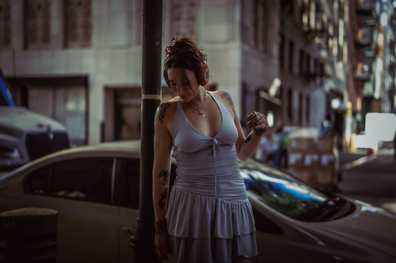 Rebekka Salomea leaning against a dark lamp post on a city street, eyes lowered, wearing red crocheted headphones over pinned-up dark hair and a pale lavender halter dress, parked cars and building fronts blurred behind her — colour portrait