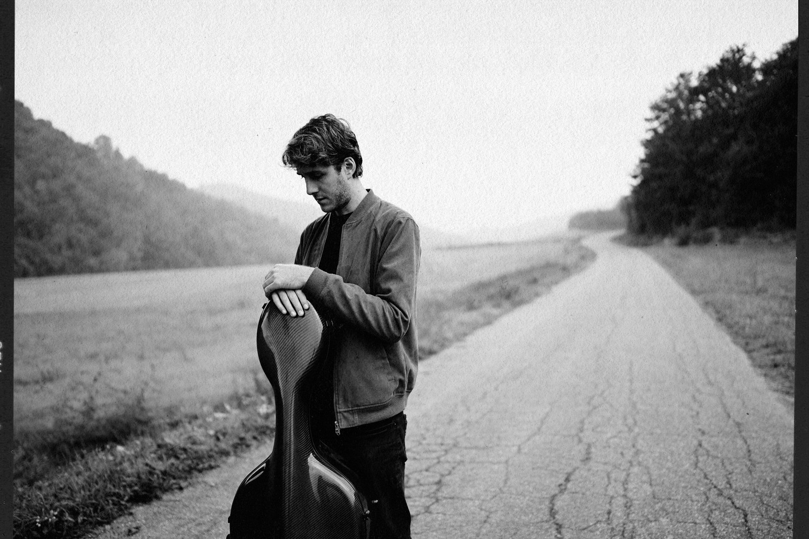Sebastian Plano on an empty country road, holding his cello case, head bowed — the visual register of Solo