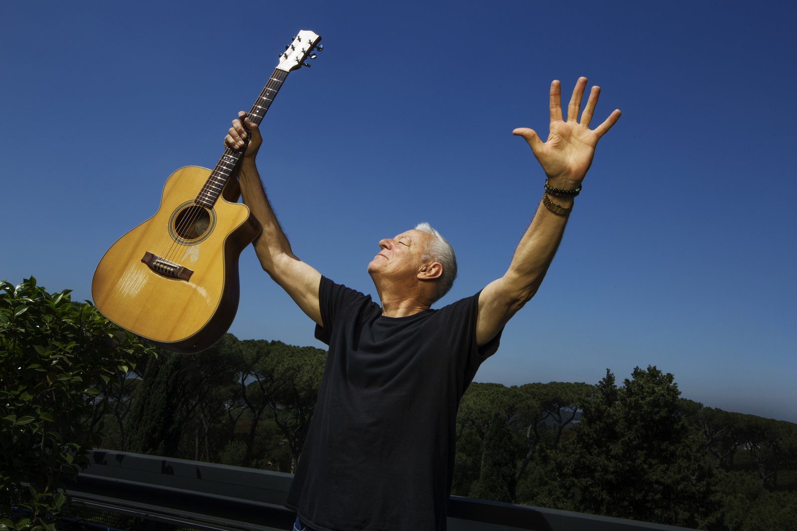 Tommy Emmanuel im Freien vor tiefblauem Himmel, die Augen geschlossen und das Gesicht erhoben, beide Arme hochgereckt — in der einen Hand seine Maton-Akustikgitarre am Hals, die andere Handfläche offen