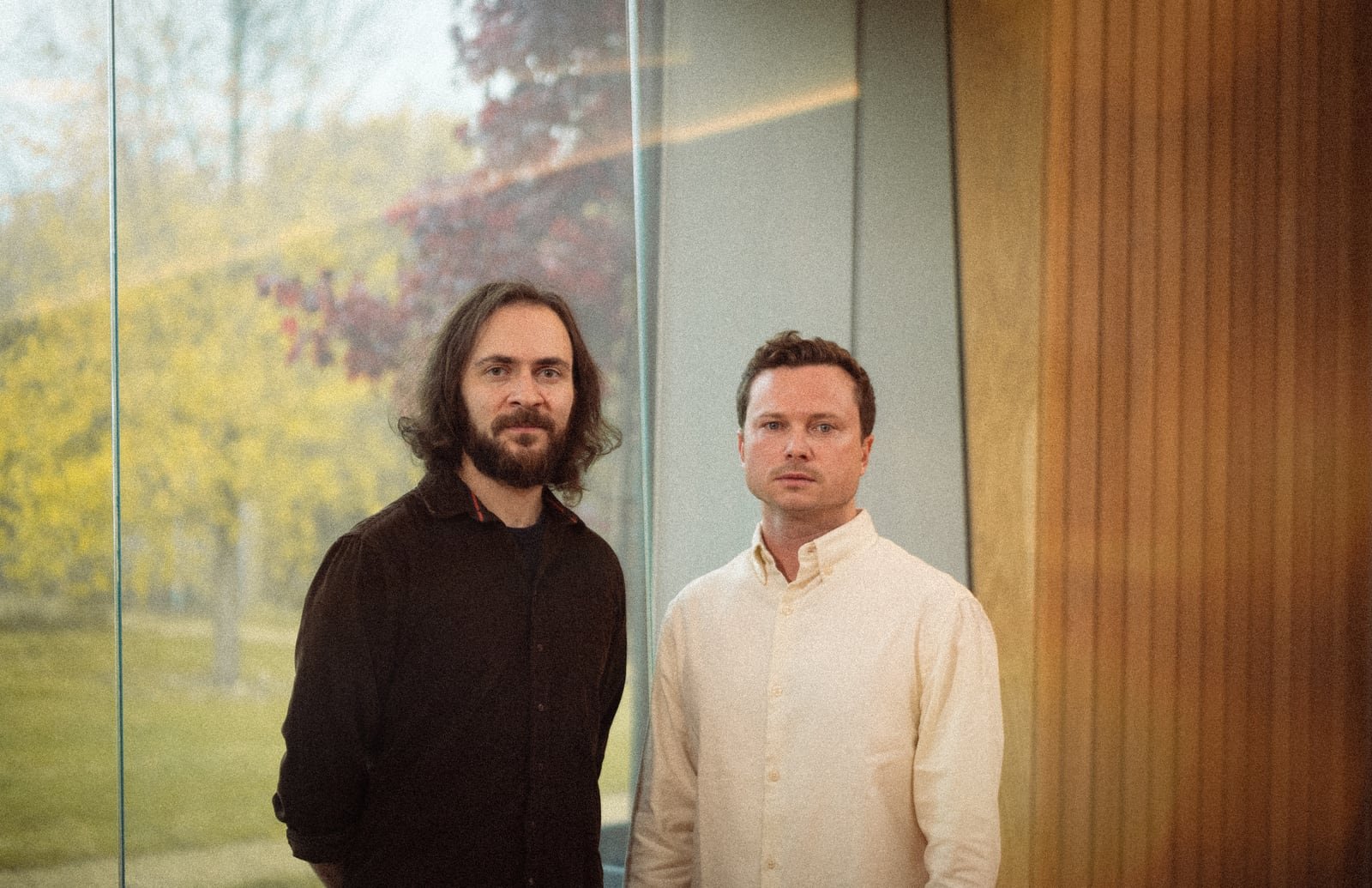 The two members of Vega Trails standing side by side against a glass wall, one long-haired and bearded in a dark shirt, the other clean-shaven in a pale cream shirt, with autumn trees beyond the glass